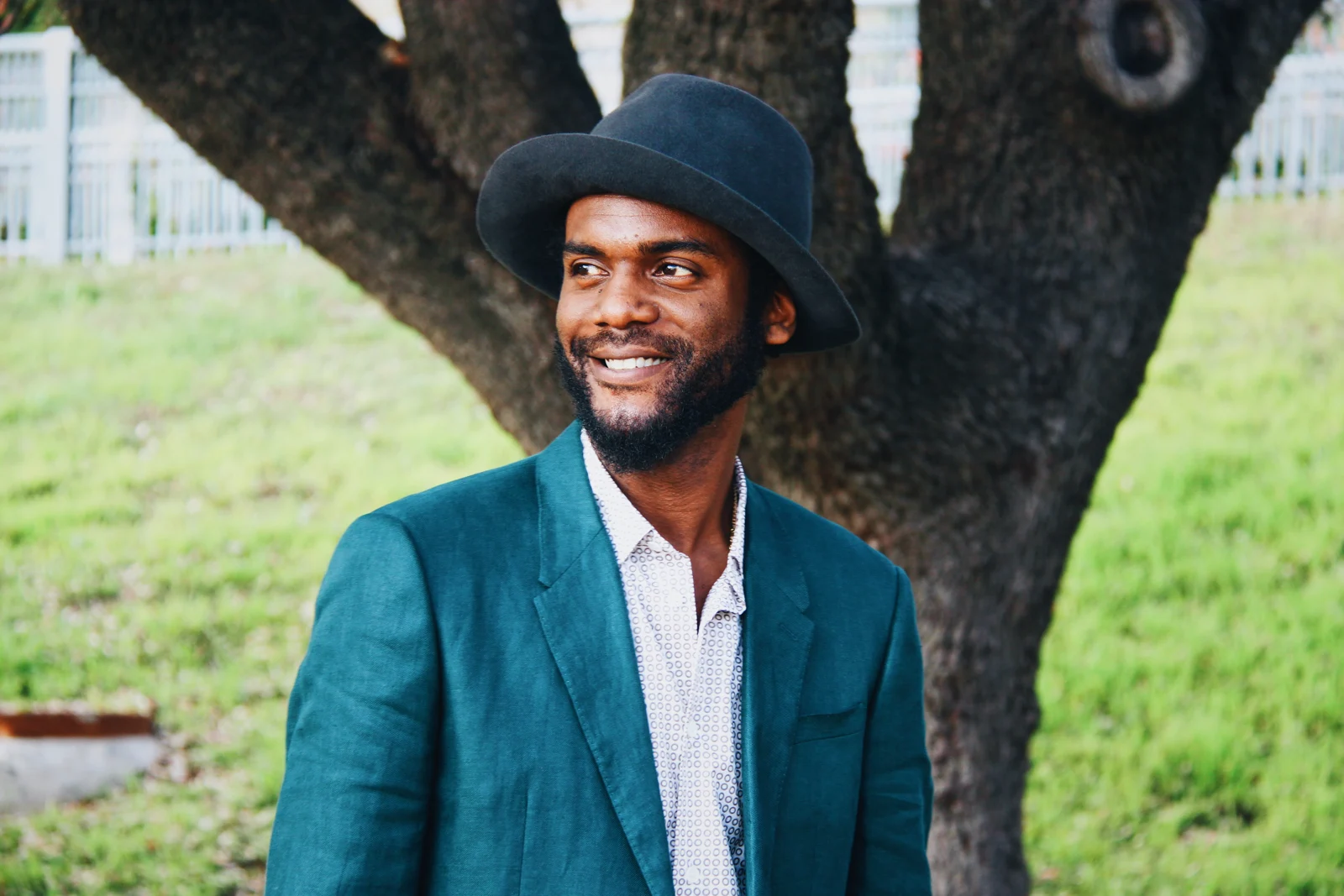 A smiling man in a teal jacket and dark hat beneath a tree